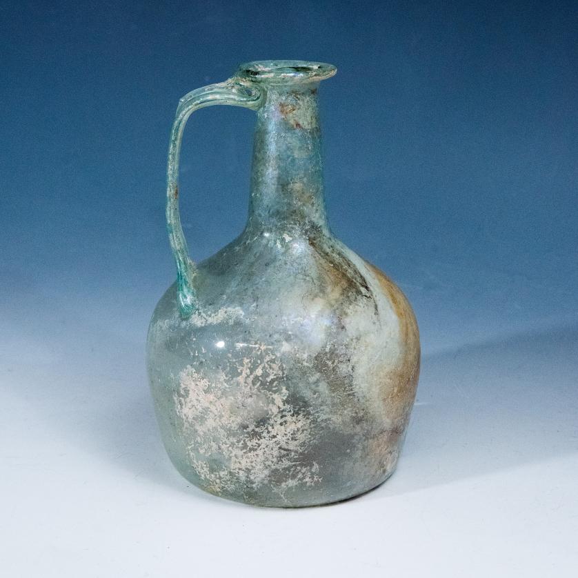 2071   -  ARQUEOLOGÍA. ROMA. Imperio Romano. Botella con asa (ss. II-III d.C.). Vidrio. Presenta irisaciones. Altura 14 cm.